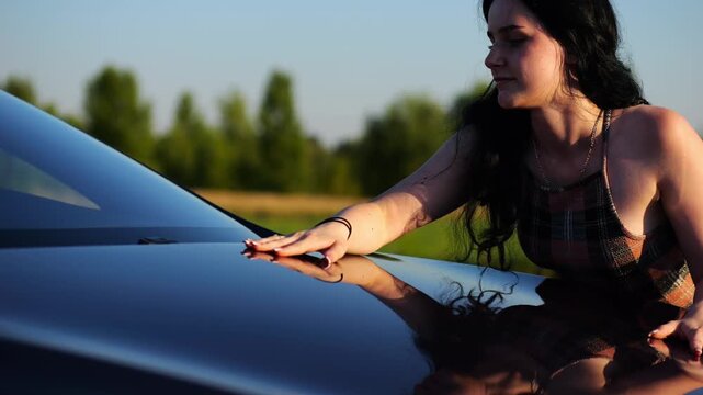 Beautiful smiling girl bought new electrical vehicle and hugging hood auto. Happy young woman enjoying modern electric automobile. Concept of buy or rent car. Slow motion