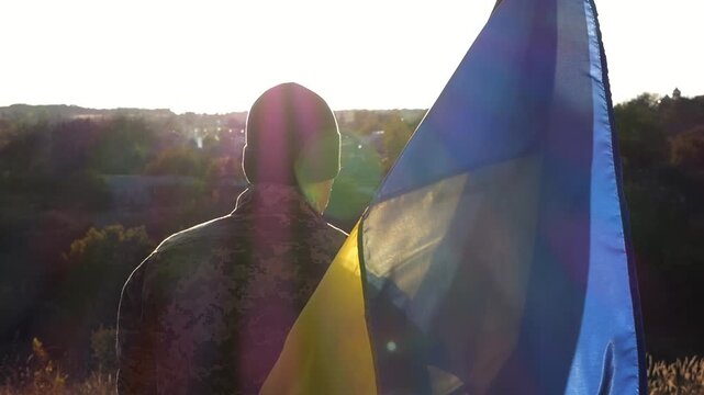 Male soldier in military uniform with Ukraine flag as symbol of victory against russian aggression. Ukrainian army man stands with national banner at countryside. Concept of invasion resistance