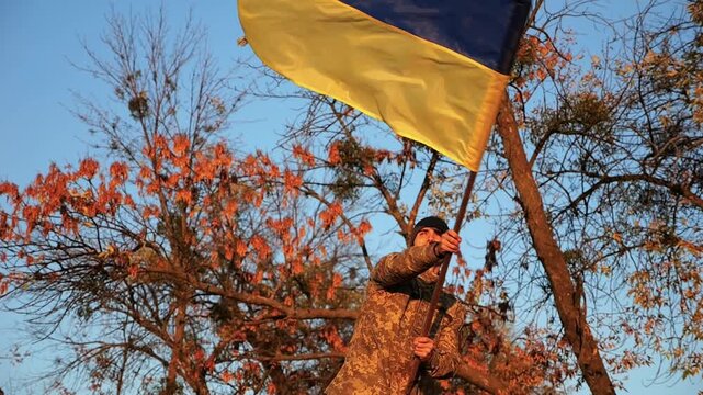 Male ukrainian army soldier waving flag of Ukraine to peak of hill at sunset. Young man in military uniform lifted national banner at countryside. Concept of victory against russian aggression