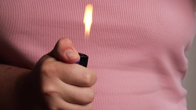 hand igniting blue plastic lighter with thumb, close up of fingers pressing flint wheel against pink knitted fabric background, faceless everyday object detail, concept of smoking habit, camping gear,