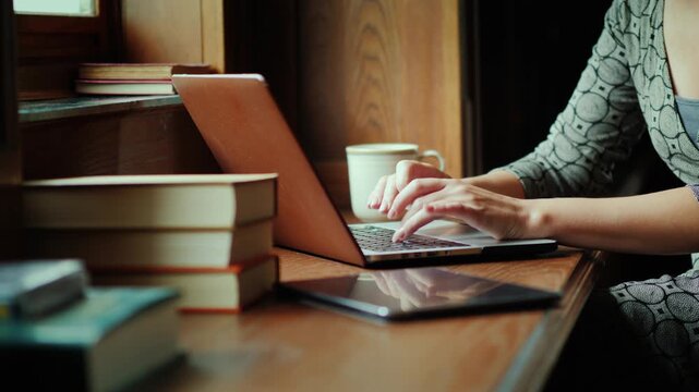 Woman typing on a laptop computer while sitting by a window in a cozy study room with stacked books and a digital tablet.