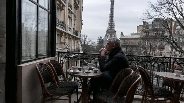 Paris, France, Europe. A man sits at a table in an outdoor cafe overlooking the Eiffel Tower in Paris, France. He is dressed in a dark jacket and appears contemplative.