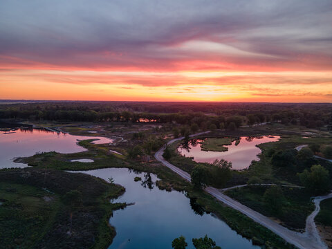 Aerial view of a winding road cutting through a landscape of reflective ponds and lush trees under a vibrant sunset sky in the Netherlands.