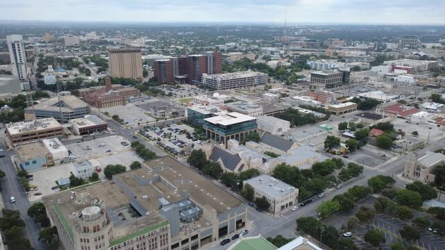 Travis Park anchors historic commercial blocks within downtown district adjacent to municipal structures. Vehicular traffic navigates grid streets beneath overcast skies in San Antonio, TX