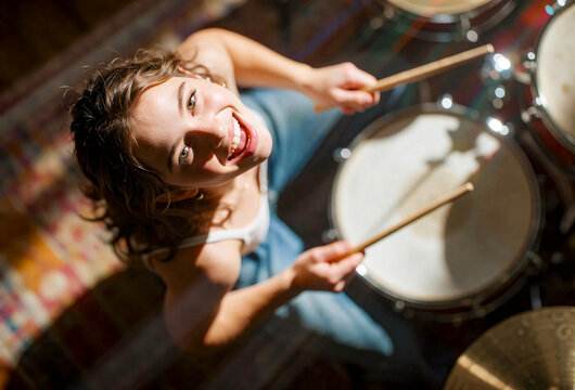 Young woman playing drums with drumsticks; sunlit indoor setting featuring wooden snare and cymbal, blue top, curly hair, overhead angle shot.