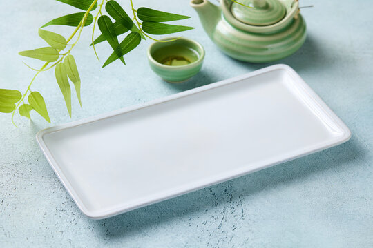 Empty white rectangular plate with green teapot and bamboo leaves. Blue light background. Close up.