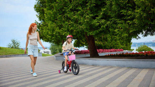 Mother supervises daughter riding bicycle independently across park plaza. Concept of growing independence, patient supervision, everyday active mobility.