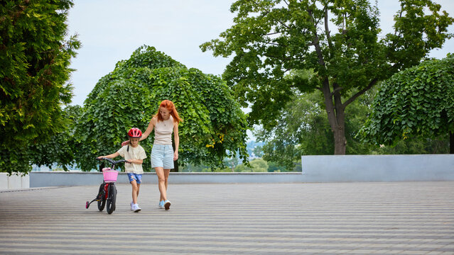 Mother walks beside daughter cycling through spacious public park. Concept of sustainable family mobility, public space recreation, healthy childhood.