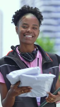 Vertical video. A delivery worker stands in the city holding packages. Busy buildings fill the background as she smiles while preparing for her next delivery. The day looks sunny.