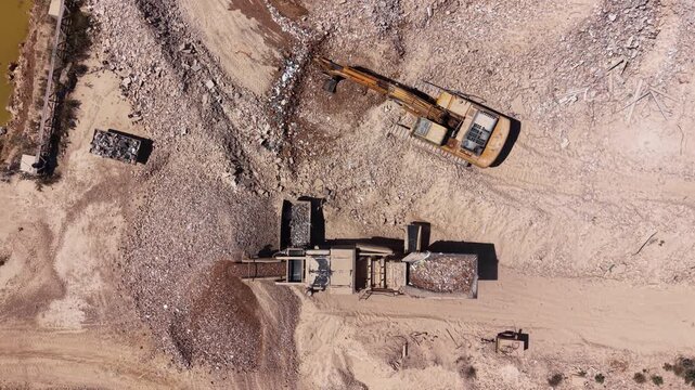 Top-down nadir view of heavy machinery including an excavator and materials handler working through piles of demolition rubble and sorted scrap at an industrial recycling site