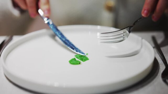 Diet concept video showing hands cutting single small green leaf on white plate with silver cutlery.