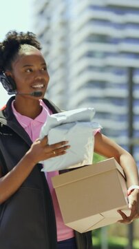Vertical video of a delivery worker walks through a busy city area carrying packages. Tall buildings surround her as she goes about her work in the bright daylight.