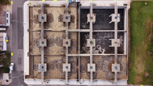 Top-down nadir view of a wastewater treatment facility showing a grid of square aeration and settling tanks with foam, sediment, and dark water patterns, bordered by road and grass