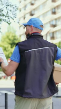 Vertical video of a delivery person checks a clipboard while walking along a busy city street. Tall buildings are in the background. Trees line the sidewalk on a sunny day.