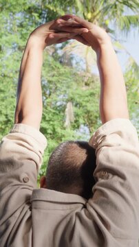 Young man performing overhead arm stretch with interlocked fingers during a peaceful morning outdoor wellness routine, improving shoulder mobility, upper body flexibility, posture, healthy lifestyle