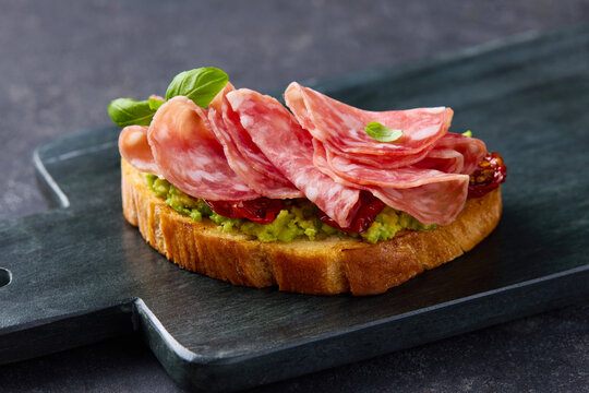 Bruschetta with salchichon, mashed avocado and sun dried tomatoes on black marble board. Dark grey background. Close up.
