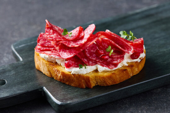 Bruschetta with salami sausage, cream cheese and pomegranate seeds on a black marble cutting board. Dark grey background. Close up.