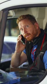 Delivery driver checks a clipboard and talks on a smartphone while seated in a parked van. Courier reviews shipping paperwork during a call, seen through the open window. Vertical video.