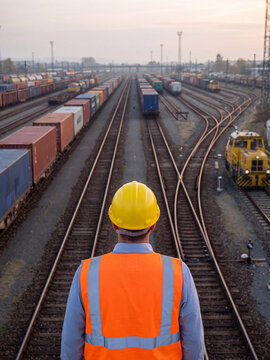 Rear view of a railway worker in a hard hat and safety vest standing in a train yard looking at freight trains