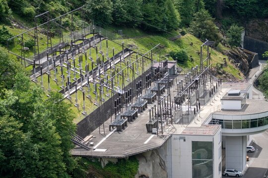 Aerial view of the Contra Dam of Verzasca Hydroelectric Power Station featuring an electrical substation with power lines on a lush green hillside in Tenero-Contra, Ticino, Switzerland.