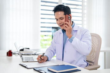 Obraz premium asian male doctor reads a medical report on a clipboard while talking on the phone in a hospital office. TeleHealth