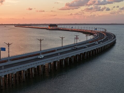 Aerial view of the Mandara Toll Road at sunset, featuring the Nusa Dua Toll Plaza with cars driving along the bridge over the calm sea in Kabupaten Badung, Bali, Indonesia.