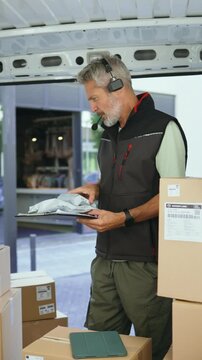 Vertical video of a delivery worker is inside a van surrounded by boxes. He checks items on a clipboard while preparing to deliver packages. The setting is an urban area with nearby buildings.
