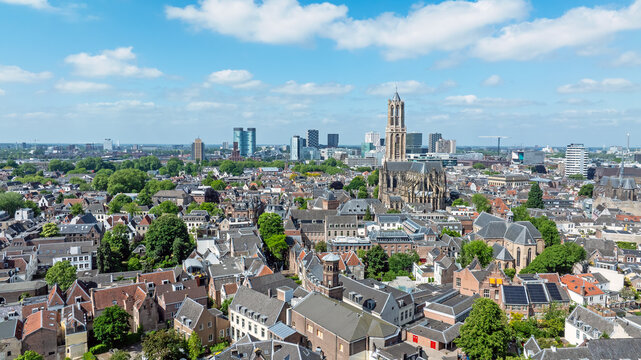 Aerial view of the historic Dom Tower rising above the dense urban landscape, featuring traditional rooftops, lush green trees, and a clear blue sky in Utrecht, Utrecht, Netherlands.