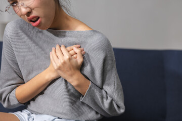 Woman clutching chest experiencing severe pain suffering from heart attack sitting on sofa indoors...
