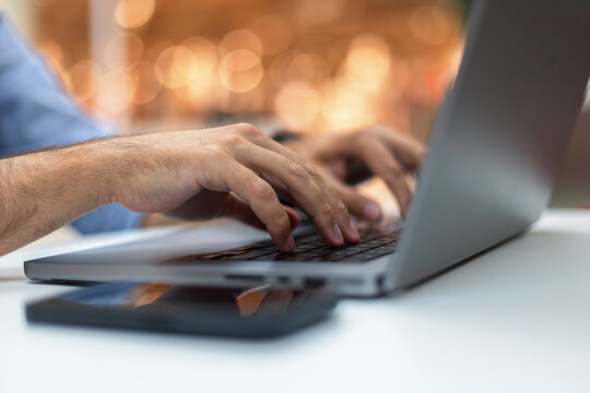 Close-up of a man's hands typing on a laptop keyboard at a desk, with a smartphone in the foreground and warm bokeh background. Perfect for freelance, coding, and remote work concepts.