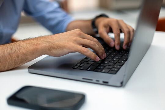 Close-up of a man's hands typing on a laptop keyboard at a white office desk, with a smartphone in the foreground. Perfect for remote work, programming, and freelance business concepts.