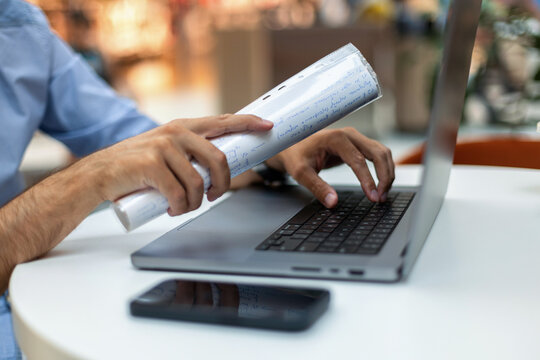 Close-up of a man's hands typing on a laptop while holding a rolled paper document at a white desk, with a smartphone in the foreground. Perfect for business, education, and planning concepts.