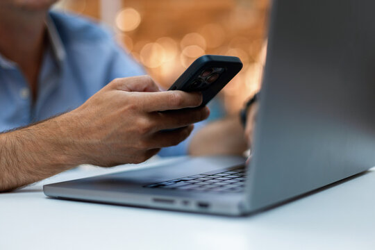 Close-up of a man's hands multitasking with a smartphone and laptop at a desk, featuring a blurred background with warm bokeh lights. Perfect for technology, remote work, and business concepts.