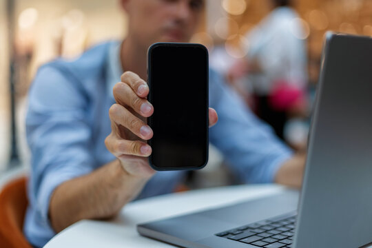Close-up of a man holding a smartphone with a blank black screen toward the camera, sitting at a desk with a laptop. Ideal mockup for mobile app, digital business, or advertisement.