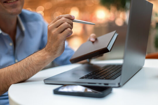 Smiling man holding a pen and notebook while working on a laptop computer at a desk. Close-up of hands planning with smartphone in foreground and warm bokeh background. Great for business concepts.