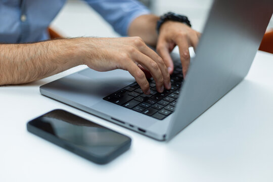 Close-up of a man's hands typing on a laptop keyboard at a white desk, with a smartphone in the foreground. Ideal for remote work, programming, business, and modern technology concepts.