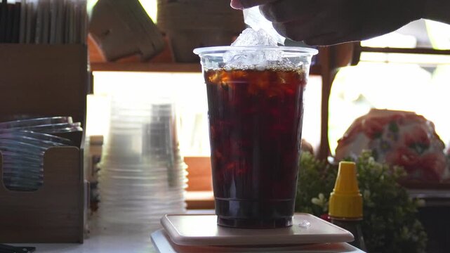 4K Barista adding extra ice cubes to cup of iced black coffee at cozy cafe counter, Concept for iced americano coffee preparation summer refreshment and cafe culture.