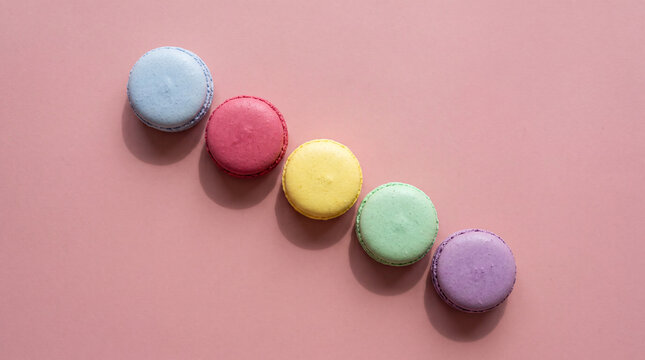Minimalist flat lay of five multicolored French macarons in a clean diagonal line over a smooth pink surface, appetizing confectionery branding design, creative food header template.