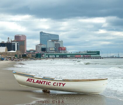 A white lifeboat labeled Atlantic City rests on a wet sandy beach with waves, a distant casino skyline and pier under dramatic cloudy skies, conveying coastal atmosphere, travel and quiet anticipation