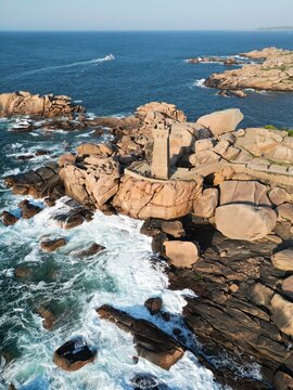 Aerial view of the Phare de Mean Ruz lighthouse on the pink granite coast, surrounded by large boulders, crashing waves, and the blue sea in Tregastel, Bretagne, France.