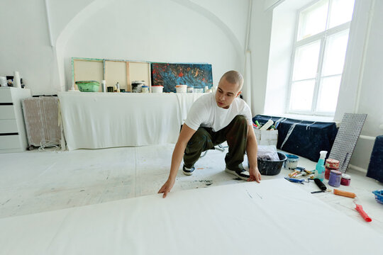 Wide shot of young adult man positioning blank canvas on studio floor, preparing contemporary artwork and creative production. Ideal for artist branding, gallery promotion, creative business