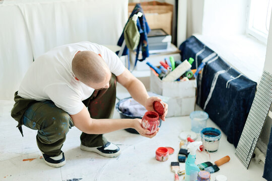 High angle view of Caucasian young adult male artist crouching and opening paint jar in studio, preparing materials for creative work. Useful for contemporary art, maker branding, workshop promotion