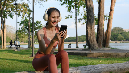 Fototapeta premium Young Asian Woman in Sportswear Using Smartphone with Headphones While Resting in Park