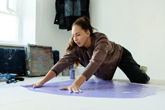 Young adult Caucasian woman spreading paint by hand on studio floor, creating contemporary artwork and expressing creativity. Ideal for art education, creative process, artist branding