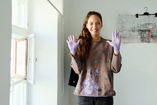 Portrait of young adult Caucasian woman smiling and showing paint covered hands in art studio, expressing creative process and artistic work. Useful for artist branding and workshop promotion