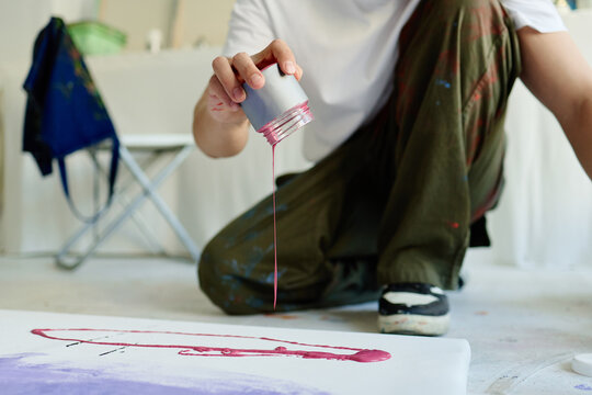 Young adult artist pouring pink paint onto canvas on floor, creating contemporary abstract artwork in studio. Ideal for creative process, modern art, artistic branding campaigns