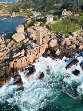 Aerial view of the Mean Ruz lighthouse on the Pink Granite Coast, surrounded by massive pink granite boulders, crashing waves, and coastal vegetation in Tregastel, Bretagne, France.
