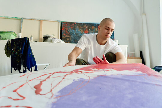 Young adult Caucasian man creating abstract painting in art studio, spreading paint by hand and developing contemporary artwork. Useful for creative business, artist branding, culture marketing