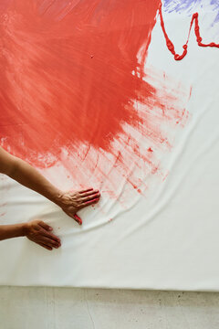 Vertical top view adult artist hands spreading red paint across large canvas, creating abstract artwork for contemporary creative process concept. Ideal for art branding, gallery promotion