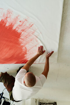 Vertical shot of middle aged man creating abstract artwork on large canvas, spreading paint by hand in studio. Ideal for contemporary art, creative process, artist branding use